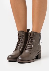 Botas de tobillo de cuero marrón oscuro con cordones, cremallera lateral y tacón bajo, llevadas sobre piernas desnudas contra un fondo blanco.