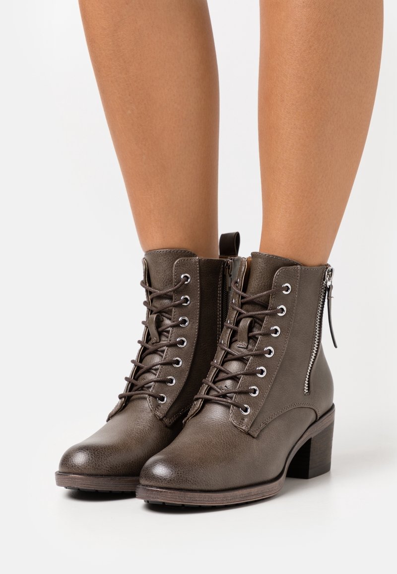 Botas de tobillo de cuero marrón oscuro con cordones, cremallera lateral y tacón bajo, llevadas sobre piernas desnudas contra un fondo blanco.