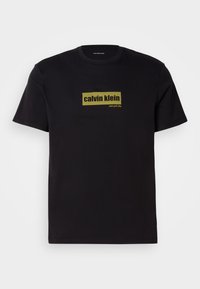 Svart bomulls-T-skjorte med korte ermer, med et rektangulært grønt logo og teksten "calvin klein" og "new york city" i fet skrift.