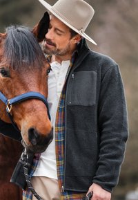 Grau gefütterte Jacke mit einer Reißverschlusstasche, über einem karierten Hemd. Das Pferd trägt eine blaue Halfter, hat einen braunen Pelz und eine kurze Mähne.