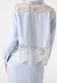 Jasnoniebieska bluzka z koronkowym panelem z tyłu, zdobiona kwiatowymi wzorami i przejrzystą teksturą. Długie rękawy i mankiety na guziki dopełniają projekt.