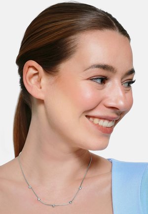 Donna sorridente con capelli castani raccolti indietro indossa una maglietta blu e una collana d'argento con piccoli ciondoli a forma di cuore.
