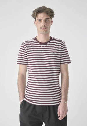 Junger Mann mit kurzem zerzaustem Haar trägt gestreiftes bordeauxrotes und weißes T-Shirt, schwarze Hose und silbernen Schmuck, steht vor einfachem Hintergrund.