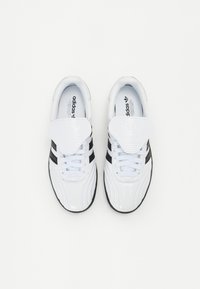 Hvite sneakers med svarte striper, overdel i syntetisk skinn, teksturert tunge, gummisåle, og synlig merking på tungen og innersålen.