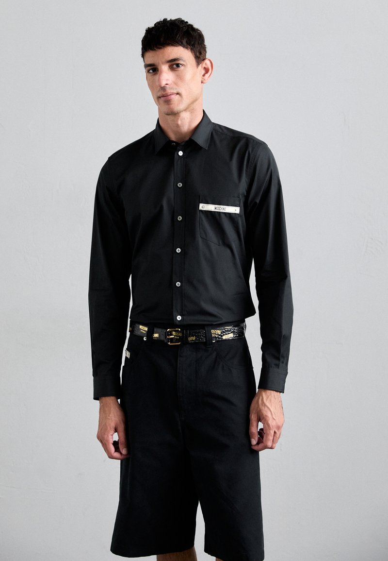 Cămașă neagră cu mâneci lungi, cu butoni, detaliu cu buzunar și accesorii din argint. Asortată cu pantaloni scurți negri și un centură lucioasă neagră și aurie.