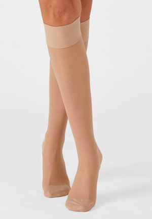 Benen met beige kniekousen van doorschijnende stof met verstevigde tenen, gekruist staand op een effen witte achtergrond.