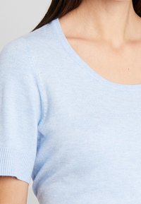 Maglione azzurro chiaro a maniche corte con scollatura rotonda e bordi a costine. Il tessuto sembra morbido con una superficie liscia e una leggera lucentezza.