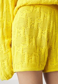 Pantalones cortos amarillos de punto con un patrón texturizado y tejidos abiertos. El diseño incluye una cintura y puños acanalados, destacando un aspecto casual.
