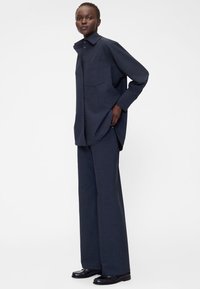 Camicia oversize blu scuro con tasca singola sul petto, abbinata a pantaloni a gamba larga dello stesso colore e scarpe nere. Tessuto liscio.