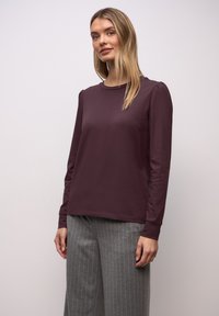 Frau mit langen blonden Haaren trägt ein dunkelviolettes Langarmoberteil und graue, gestreifte, weit geschnittene Hosen, steht vor einem schlichten hellen Hintergrund.