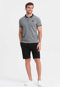 Graues Poloshirt mit schwarzem Kragen und Bündchen, schwarze Shorts und weiße Slipper. Mit einem Logo auf der Brust. Lässige Passform.