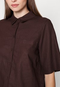 Camisa de manga curta bordô, feita de linho, com gola, bolso frontal e fecho de botões ao longo da abertura.