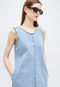 Γυναίκα με ίσια καστανά μαλλιά που φοράει μαύρα γυαλιά ηλίου Calvin Klein και ένα αμάνικο ανοιχτό μπλε τζιν φόρεμα με τσέπες, στέκεται μπροστά σε ένα λευκό φόντο.