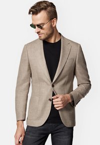 Beżowa żakiet o fakturze z dopasowanym krojem, z dwoma guzikami i kieszeniami z przodu, noszona na czarnej koszulce. Brązowe akcenty guzikowe dopełniają projekt.