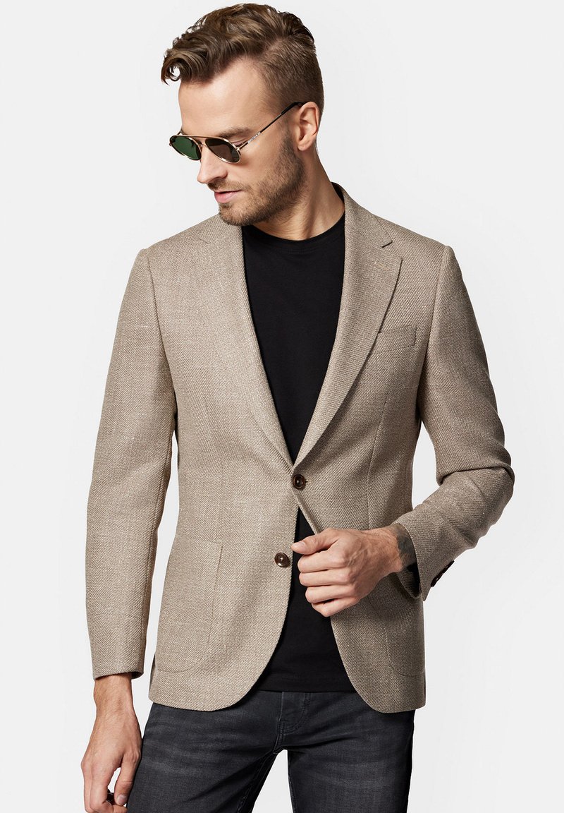 Beżowa żakiet o fakturze z dopasowanym krojem, z dwoma guzikami i kieszeniami z przodu, noszona na czarnej koszulce. Brązowe akcenty guzikowe dopełniają projekt.