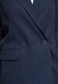 Blazer bleu marine à double boutonnage avec fines rayures blanches, deux boutons foncés et poches à rabat.