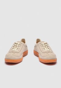 Pár béžových semišových tenisk so šnúrkami v bielej farbe a pomarančovými gumovými podrážkami, zobrazený spredu na jednoduchom pozadí.