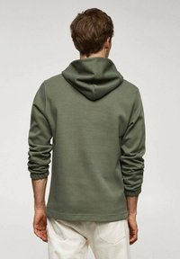 Sweat à capuche vert foncé à texture douce, avec un panneau arrière, une capuche à cordon de serrage et des poignets côtelés. Porté avec un pantalon blanc.