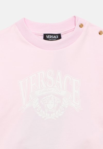 Ροζ βαμβακερό μπλουζάκι με λευκό λογότυπο Versace και κλασικό σχέδιο. Διαθέτει κουμπιά σε χρυσή απόχρωση στον ώμο και ριμπ λαιμόκοψη.