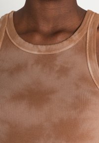 Brun ribbad tanktopp med rund halsringning och slät tyg med ett subtilt tie-dye-mönster. Accenter inkluderar kontrasterande sömnadsdetaljer.