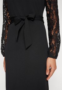 Robe noire avec des manches longues en dentelle à motifs floraux. Une ceinture en tissu nouée à la taille rehausse la silhouette de la pièce. Texture lisse.