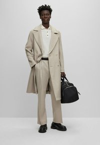 Beiger doppelt-knöpfiger Mantel, cremefarbenes Polo und weit geschnittene Hose. Schwarze Tasche mit Reißverschluss. Schwarz-weiße Schuhe mit dicker Sohle. Schlichtes, modernes Design.