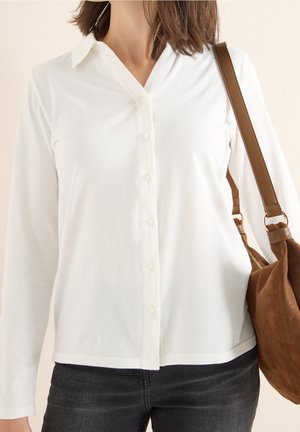 Woman wearing a white button-up long sleeve shirt with dark jeans and carrying a brown suede shoulder bag on beige background.