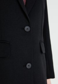 Čierny blejzer vyrobený z textúrovanej tkaniny, s dvoma prednými vreckami, jedným radom gombíkov a s límcom.