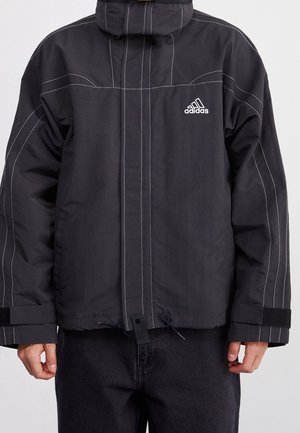 Veste noire Adidas avec coutures blanches, col montant, fermeture éclair à l'avant et poignets réglables avec velcro, portée par une personne avec un pantalon noir.