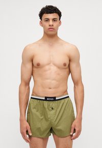 Olive green boxer shorts with an elasticated waistband featuring a contrasting black and beige logo. Lightweight fabric, loose fit.