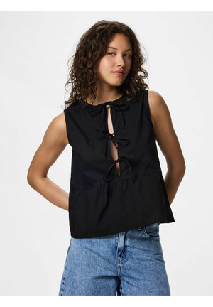 Jeune femme aux cheveux bouclés portant un haut noir sans manches avec trois nœuds à l'avant et un jean en denim bleu clair, debout devant un fond blanc.