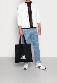 Czarna torba na ramię z logo "new balance", trzymana przez osobę w białej kurtce, w pasiastą czarną i czerwoną koszulę oraz jasnoniebieskie dżinsy. Białe sneakersy.