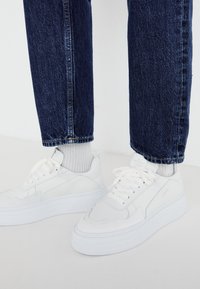 Zapatillas deportivas blancas con un diseño texturizado y suelas gruesas, combinadas con calcetines de canalé gris claro y jeans de mezclilla azul oscuro.