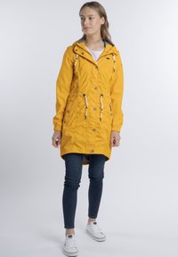 Sárga vízálló dzseki húzózsinórral ellátott kapucnival, cipzáras és patentos záródással, valamint praktikus zsebekkel. Sötét farmernadrág és fehér sportcipő kíséretében.