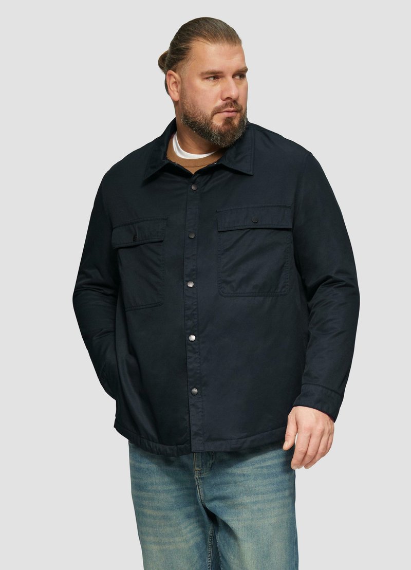 Marineblauwe jas met knoopsluiting, twee borstzakken en een kraag. Draagt over een beige shirt, gecombineerd met lichtblauwe jeans.