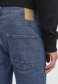 Modre kavbojke iz jeansa z teksturirano površino, ki imajo na zadnjem delu pasu rjav usnjen dodatek ter en zadnji žep z natančnimi šivi.