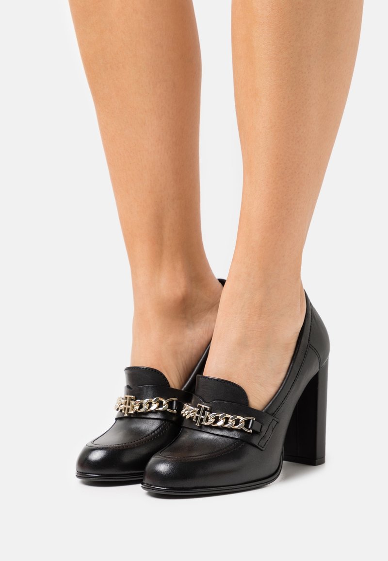 Mocassins en cuir noir à talons hauts avec détail de chaîne dorée portés pieds nus, debout sur un fond clair uni.