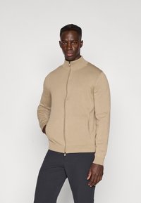 Πουλόβερ μπεζ με φερμουάρ, υψηλός γιακάς και μακριά μανίκια, με λεία υφή και εφαρμοστή σχεδίαση· σε συνδυασμό με σκούρο παντελόνι.