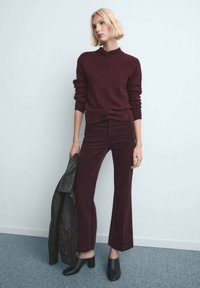 Femeie cu păr scurt blond, purtând un pulover burgundy și pantaloni asortați, ținând o jachetă de piele neagră și purtând pantofi negri cu toc.