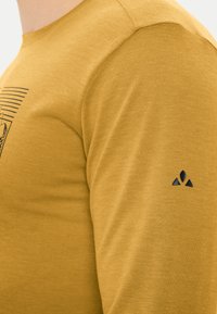 Chemise à manches longues de couleur moutarde, en tissu doux, ornée d'un design graphique sur le devant et d'un logo noir sur la manche gauche.