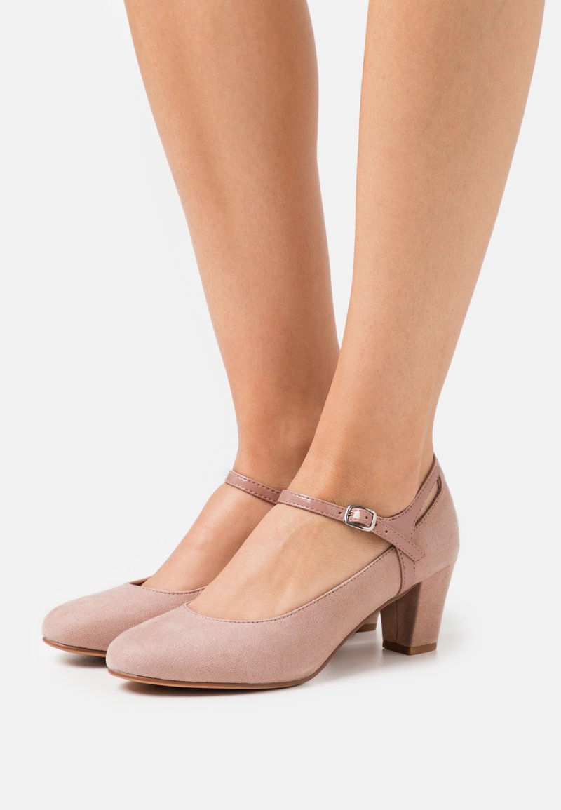 Pieds d'une femme portant des escarpins Mary Jane en suède rose clair avec bride ajustable à la cheville et talon bloc sur fond blanc.