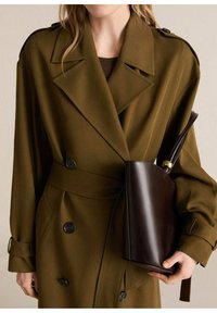 Trench coat de color verde oliva, de doble botonadura, con hombreras, bolsillos laterales y un cinturón a juego. Bolso de piel marrón con un acento dorado.