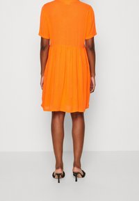 Oranges kurzärmliges Kleid mit einem ausgestellt Rock. Leichter Stoff; schlichtes Design mit rundem Ausschnitt und lockerer Passform. Schwarze Hochzeitsandalen.