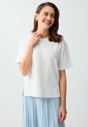 Uśmiechnięta kobieta o brązowych włosach, ubrana w biały top z krótkim rękawem i jasnoniebieską plisowaną spódnicę, dotykająca dekoltu, na jednolitym szarym tle.