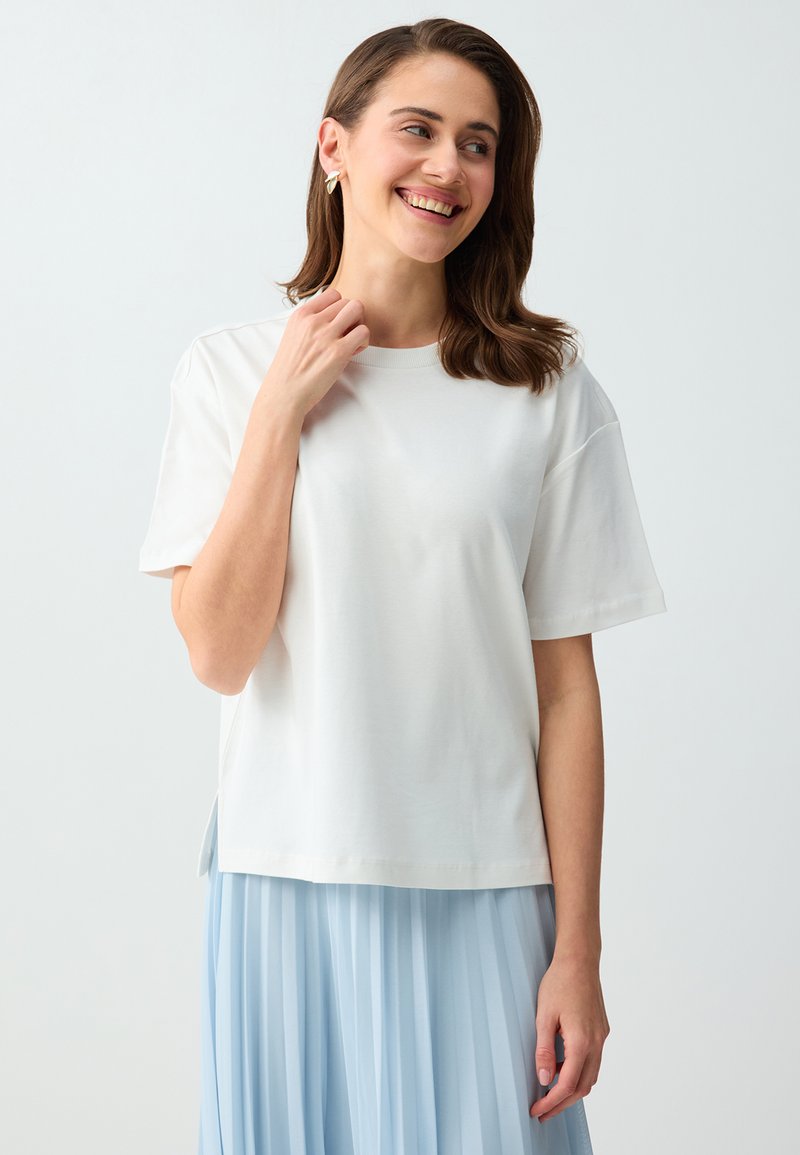 Femme souriante aux cheveux bruns portant un haut blanc à manches courtes et une jupe plissée bleu clair, touchant l'encolure, sur un fond gris uni.