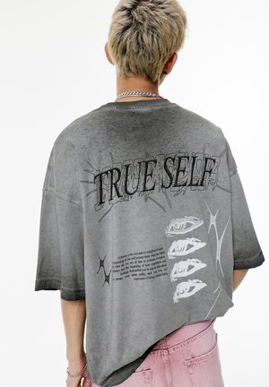 Person with short blond hair wearing a gray oversized T-shirt with graphic text "TRUE SELF" and abstract designs on the back, paired with pink pants.