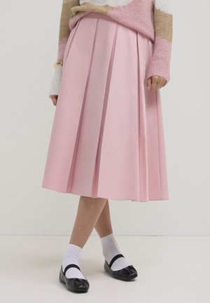 Plissierter Rock aus pastellrosa Stoff, knielang mit glatter Textur. Kombiniert mit weißen Socken und schwarzen flachen Schuhen.