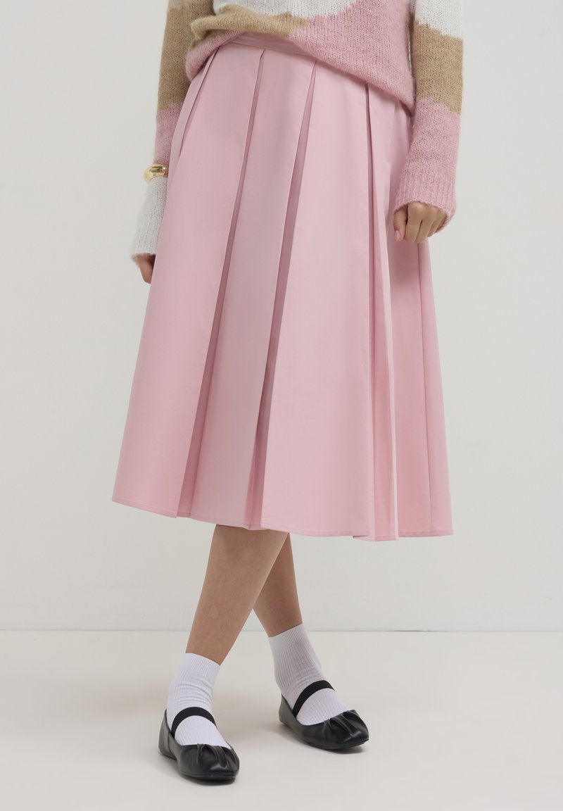 Gonna plissettata in tessuto rosa pastello, lunghezza ginocchio con una texture liscia. Abbinata a calze bianche e scarpe basse nere.