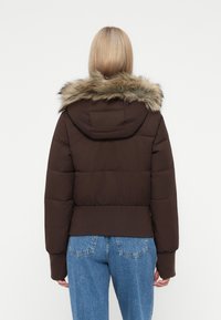 Žena s blond vlasy, oblékající hnědou prošívanou bundu s kapucí s kožešinovým lemem a modré džíny, stojící zády k fotoaparátu na bílém pozadí.