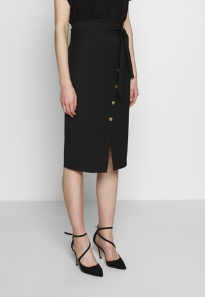 Frau trägt einen schwarzen knielangen Rock mit vorderen Knöpfen und seitlichem Schlitz, kombiniert mit schwarzen spitzen Pumps mit Knöchelriemen.